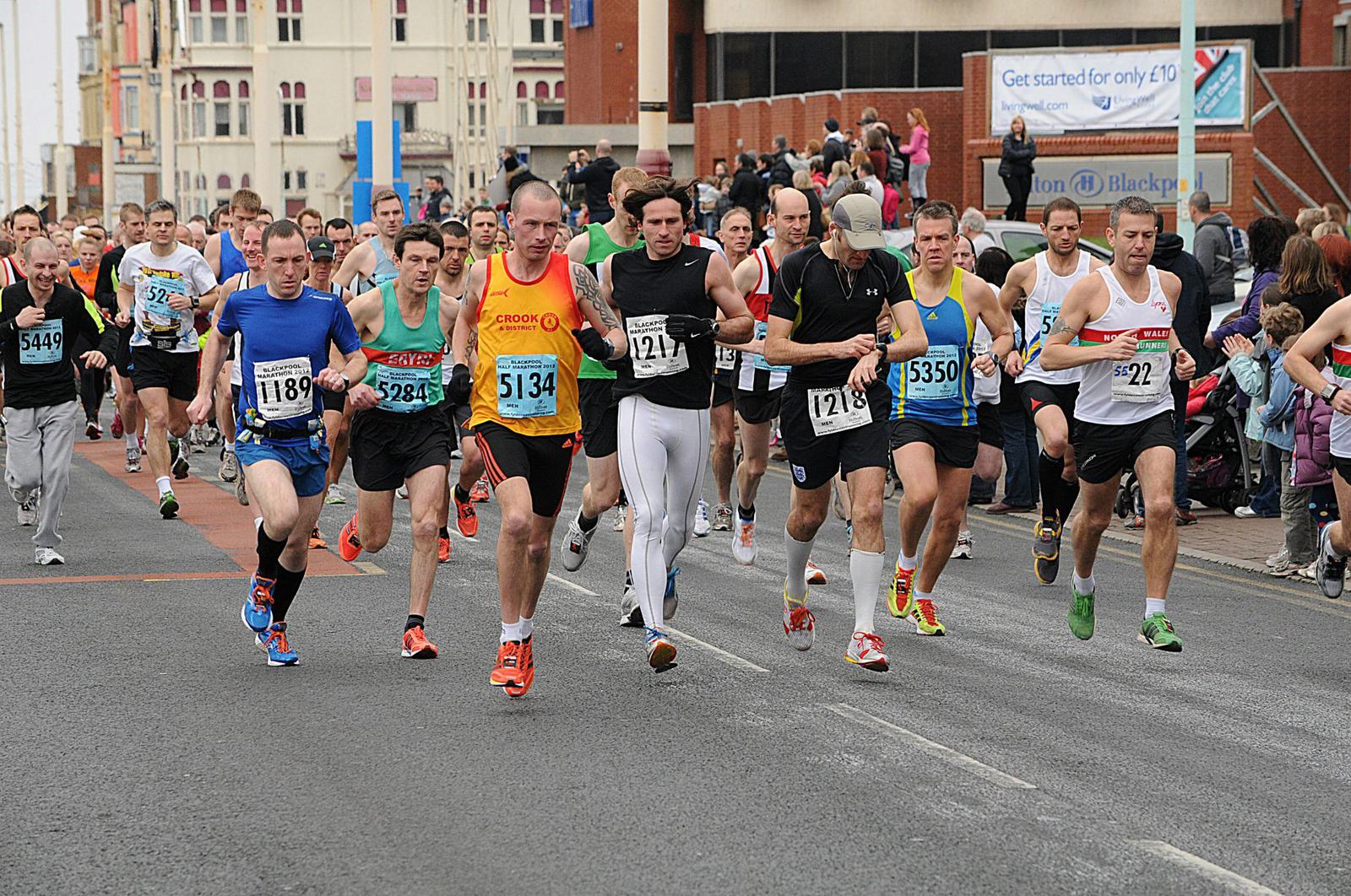 A group of runners in numbered bibs competes in a marathon on a city street. The diverse crowd of athletes is in mid-stride, with spectators lining the side of the road. Buildings and banners are visible in the background.