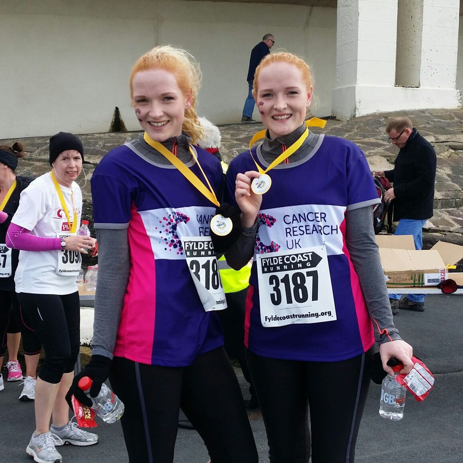 Two women wearing race numbers and medals smile at the camera. They're dressed in matching athletic gear with "Cancer Research UK" logos. Other runners and bystanders are visible in the background.