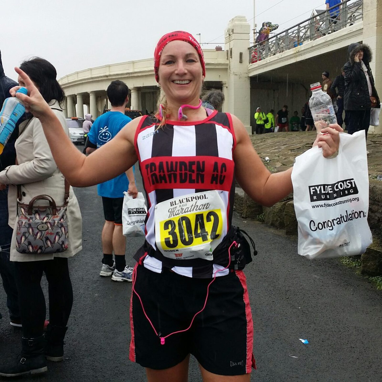 A smiling runner wearing a "Trawden AC" jersey and the race number 3042 holds a water bottle and goodie bag. They stand on a wet path near a white building, surrounded by other participants and spectators.
