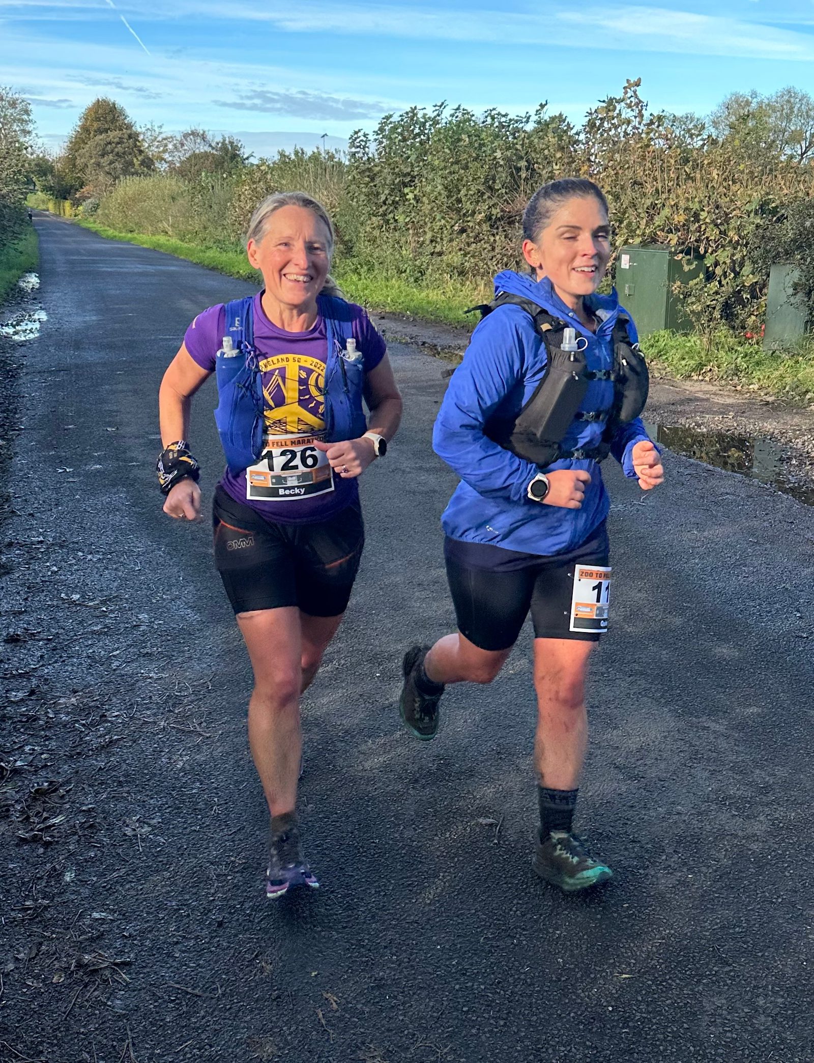 Two women running on a rural path, wearing race bibs and hydration packs. One wears a purple and yellow top, and the other wears a blue jacket. It's a sunny day with greenery in the background. Both are smiling and appear to be in a race or marathon.
