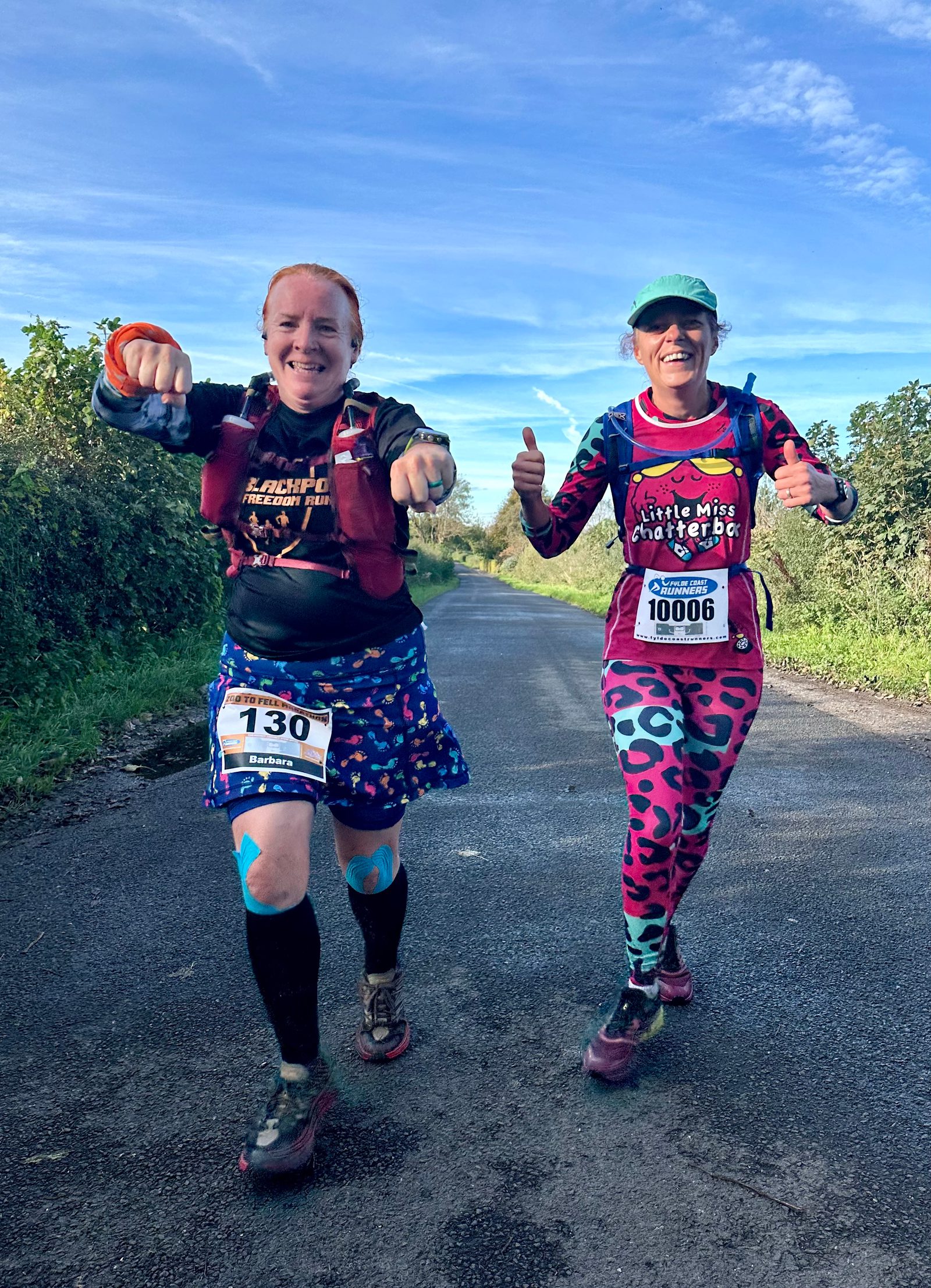 Two marathon runners smile and gesture towards the camera on a sunny day. They wear colorful outfits and race bibs numbered 130 and 1006. The road is surrounded by greenery, and the sky is clear and blue.