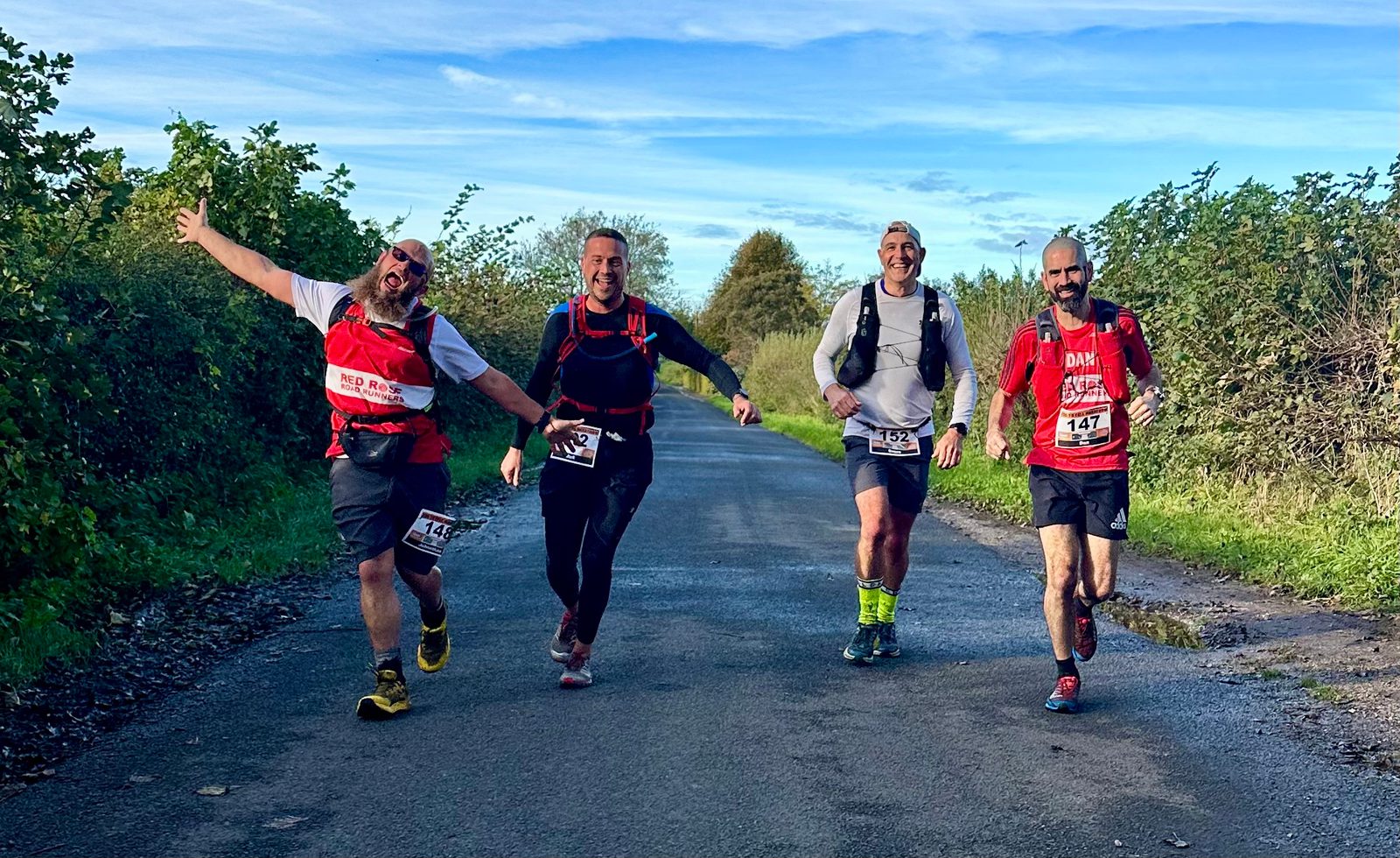 Four men are jogging on a rural road under a blue sky. They appear joyful, with one man playfully raising his arms. They wear running gear, including numbered bibs and backpacks, surrounded by green bushes and trees.