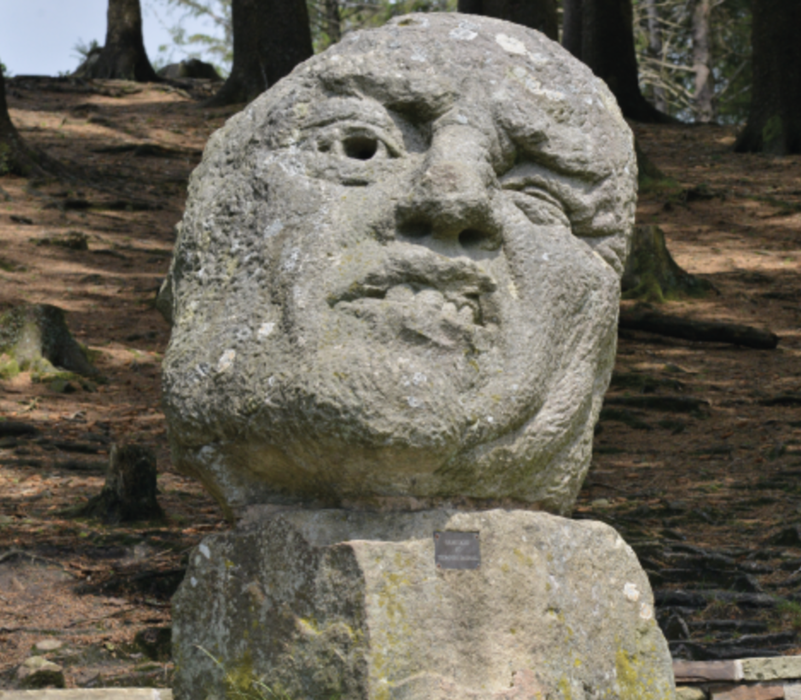 A large, weathered stone sculpture of a human face with exaggerated features is set in a forested area, with trees and dappled sunlight in the background. The sculpture rests on a stone pedestal.