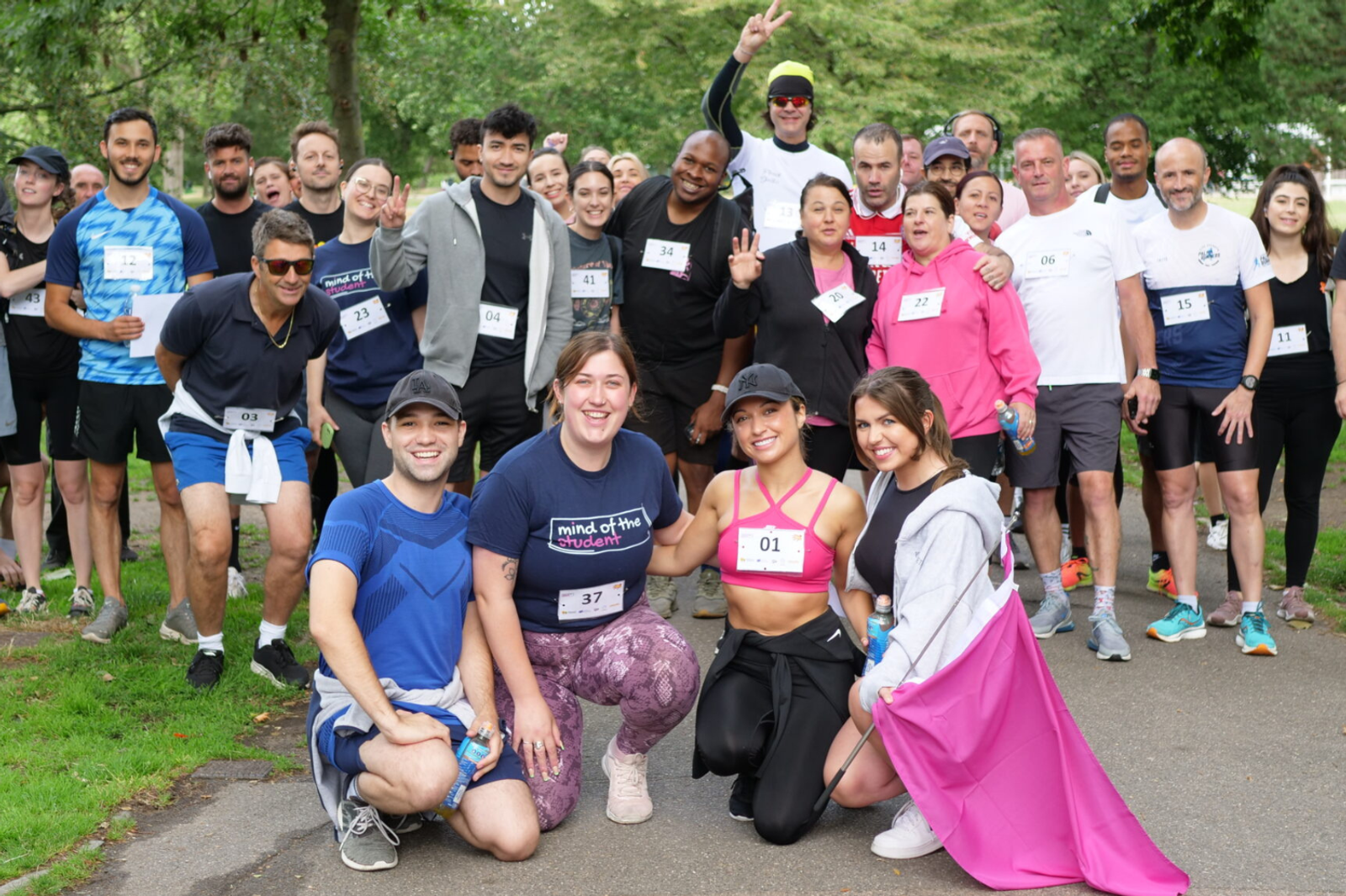 A diverse group of runners stands together in a park, smiling and posing for a photo. Some wear numbered bibs while others hold up peace signs or wave. Trees and greenery are visible in the background, suggesting a vibrant, outdoor event or race.