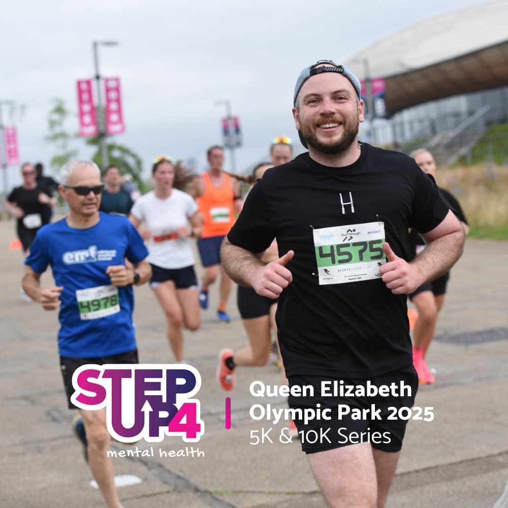 A group of smiling runners participate in a race at Queen Elizabeth Olympic Park. One man in front, wearing black, holds his bib number 4375. Event banners and the STEP UP 4 mental health logo are visible.