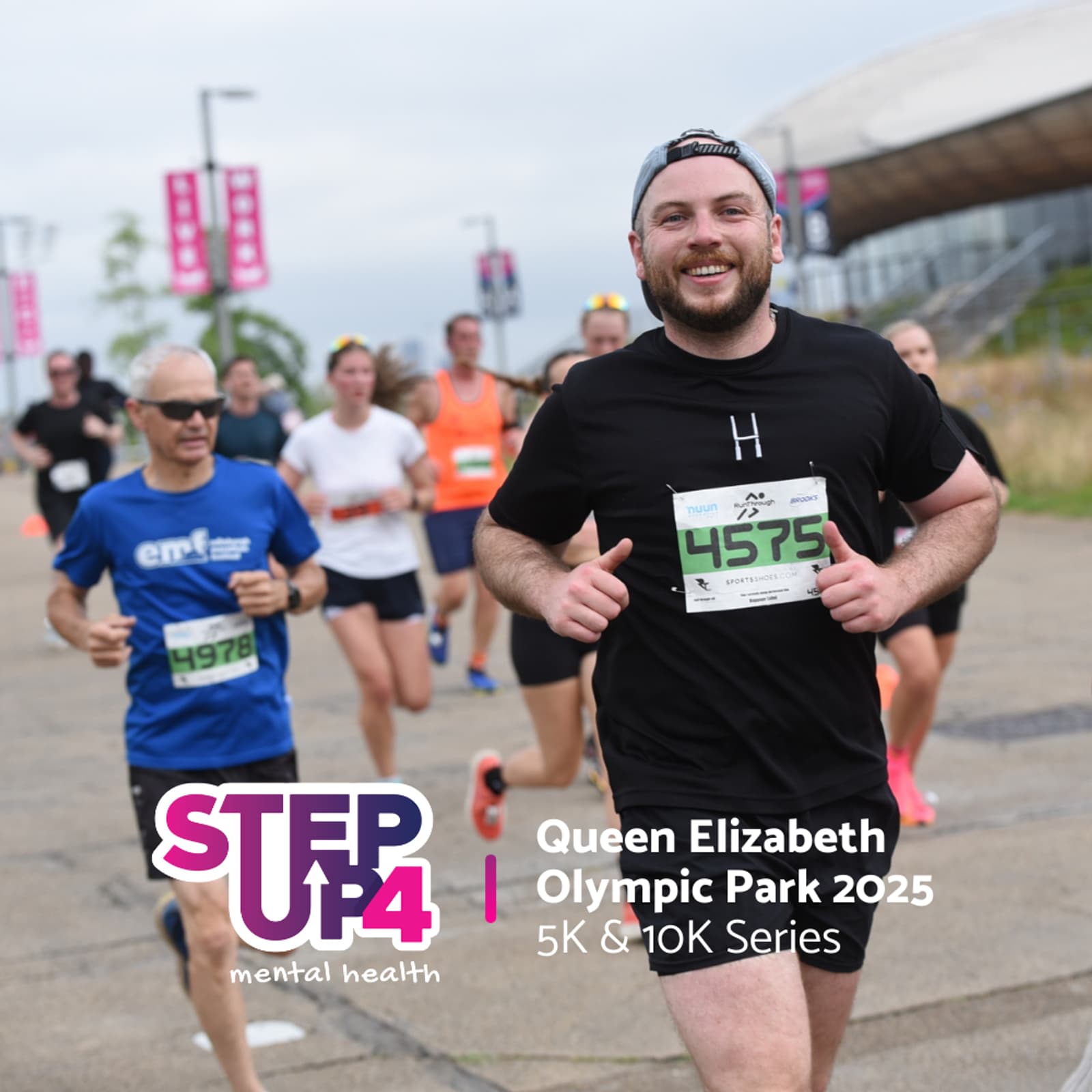 A group of smiling runners participate in a race at Queen Elizabeth Olympic Park. One man in front, wearing black, holds his bib number 4375. Event banners and the STEP UP 4 mental health logo are visible.