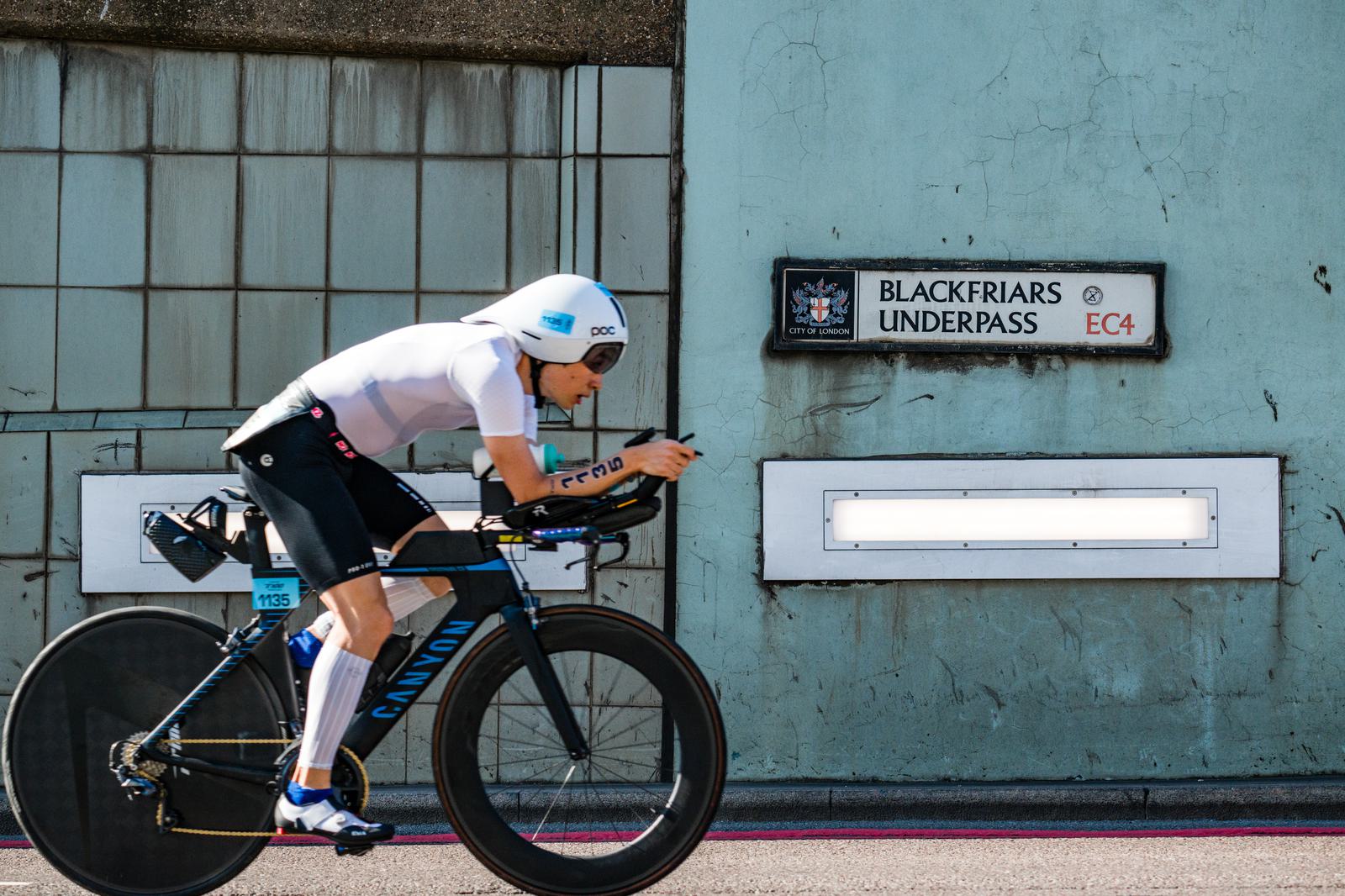 Cyclist in aerodynamic gear rides a blue and black bike past a sign reading "Blackfriars Underpass EC4." The background features a concrete wall with evenly spaced rectangular lights.
