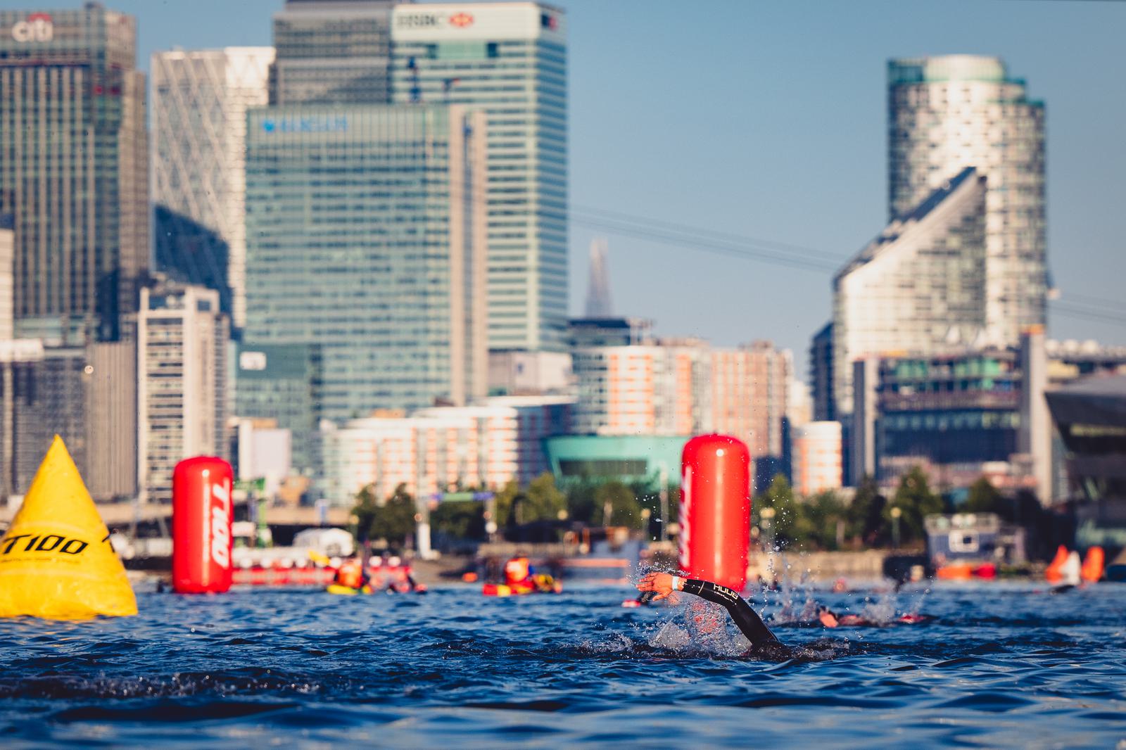 Swimmer in a wetsuit competes in a triathlon, cutting through the water with city skyscrapers in the background. Bright buoys mark the course as safety personnel in kayaks supervise the event. The sun casts a clear light on the scene, indicating good weather.