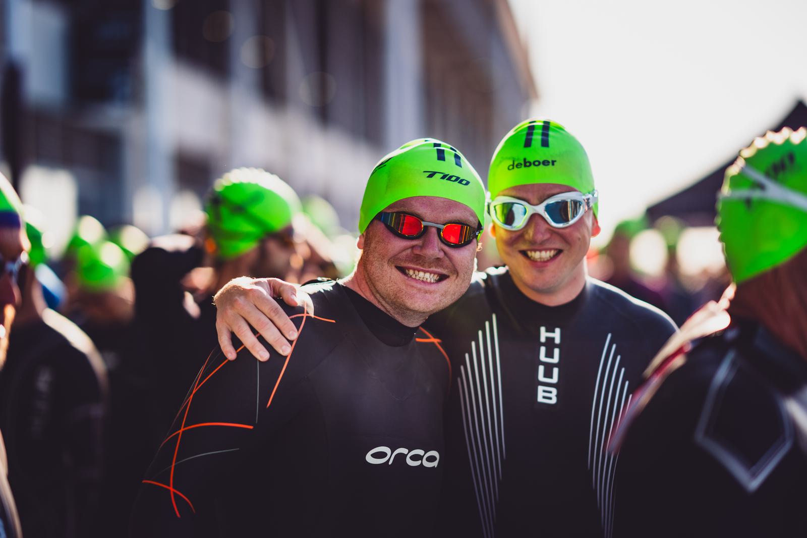 Two participants in swimming wetsuits and green swim caps smile for the camera. One has an arm around the other's shoulder. They are standing among a group of other participants who are also wearing similar swim gear. The background shows blurred structures.