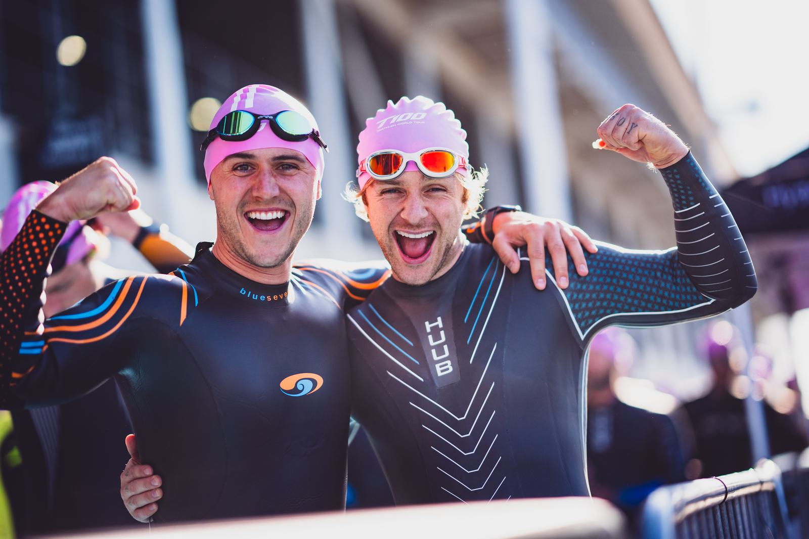 Two triathletes in wetsuits and swim caps smile and flex their biceps at a race event. They stand close together with their arms around each other’s shoulders, capturing a moment of joy and camaraderie before the competition.