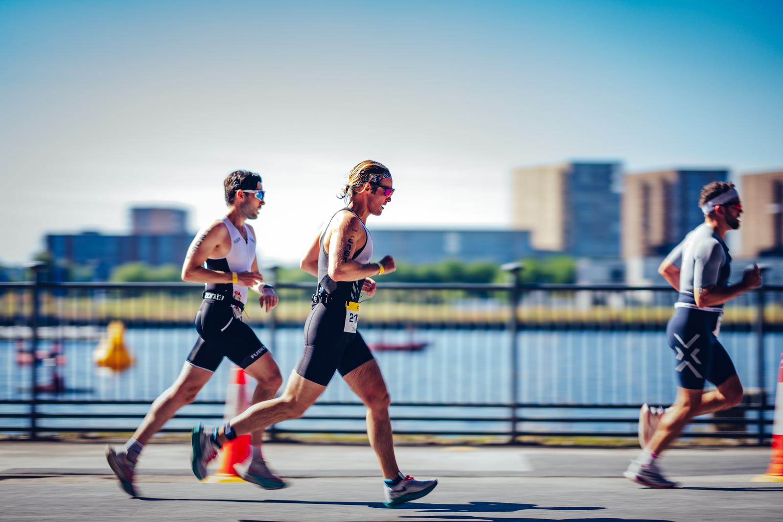 Three athletes are running on a sunny day, likely participating in a triathlon or race, wearing athletic gear and numbered bibs. They are running next to a body of water, with blurred buildings and structures in the background.