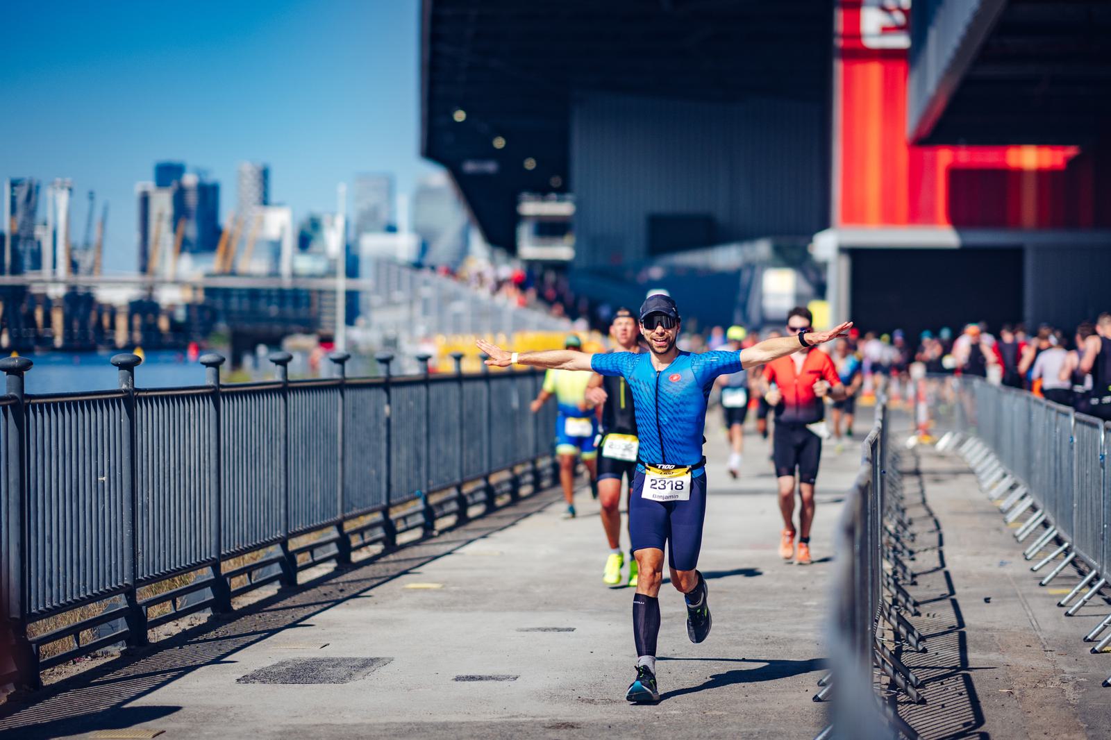 A runner dressed in blue spreads his arms wide while approaching the finish line during a race. He is wearing a race bib, sunglasses, and a cap. Other runners are visible behind him on the sunny, open course bordered by a metal fence and a cityscape in the background.