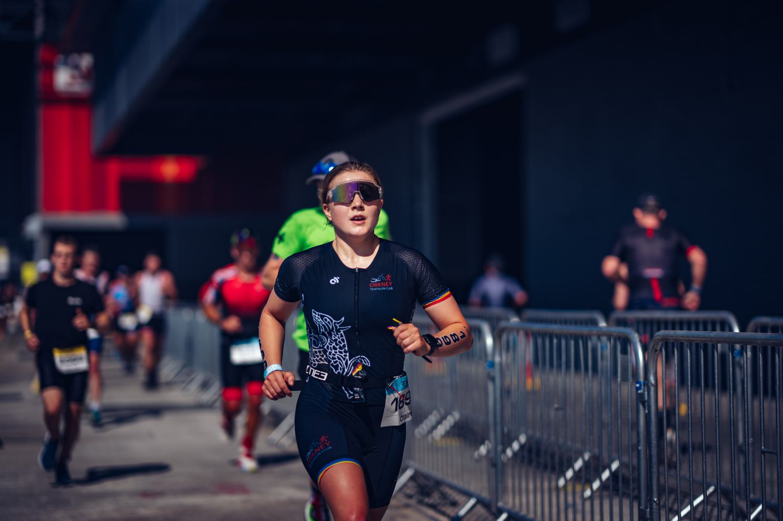 A focused participant wearing sports attire and sunglasses runs in a race with other runners visible in the background. Metal barricades line the path, and the scene takes place under a bridge or overpass. The event atmosphere appears sunny and energetic.