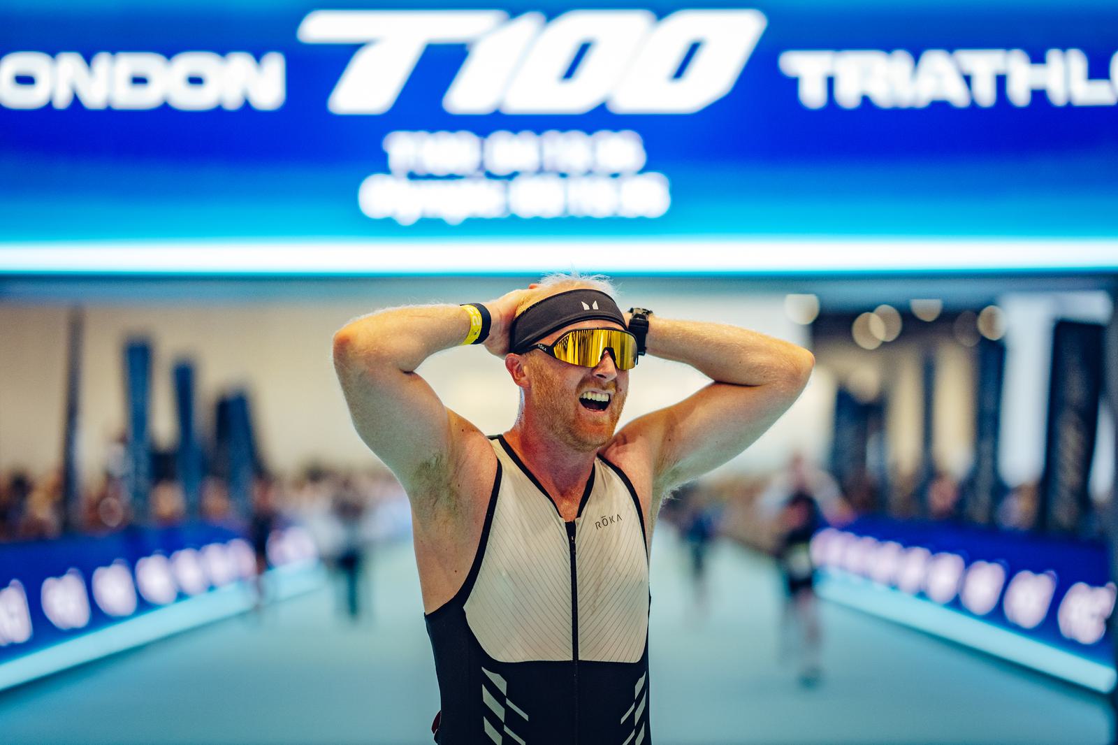 A participant in a triathlon smiles and holds his head in relief as he crosses the finish line. He is wearing a sleeveless triathlon suit, sunglasses, and a cap. A large electronic display overhead shows "London Triathlon" and the distance "100.