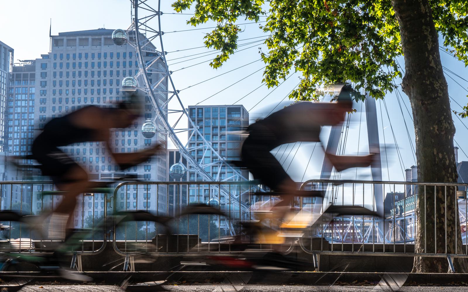 Two cyclists in motion ride past a backdrop of the London Eye and city buildings. Blurred motion captures their speed, while sunlight filters through the trees above.