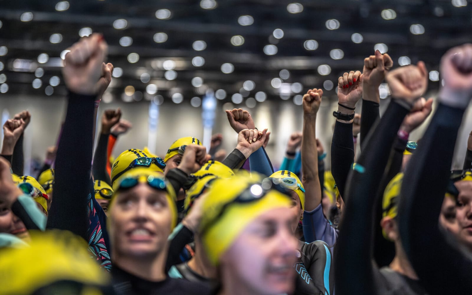 A group of swimmers in yellow swim caps and goggles raise their fists in the air, celebrating or cheering together in a brightly lit indoor setting. The image has a vibrant, energetic atmosphere.