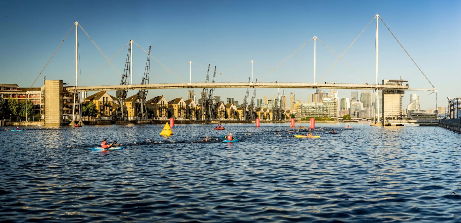 People kayak on a calm river beneath a modern suspension footbridge, with industrial cranes and unique riverside buildings in the background; a city skyline is visible under a clear blue sky.