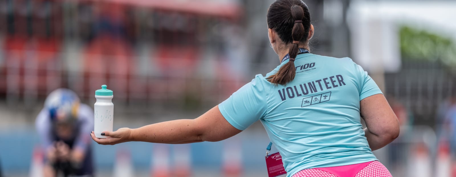 A volunteer wearing a blue shirt extends a water bottle towards an approaching athlete at an outdoor sporting event, with blurred spectators and barriers in the background.