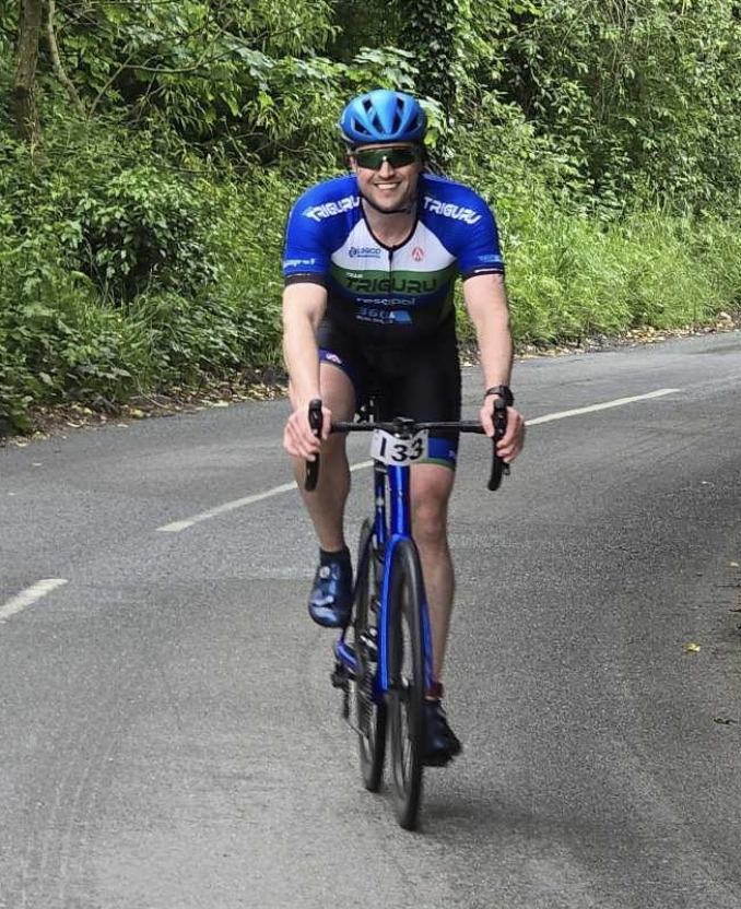 A cyclist wearing a blue helmet and blue cycling jersey, numbered 134, rides on a paved road surrounded by greenery. The cyclist is focused and appears to be participating in a race or event, with trees and foliage in the background.
