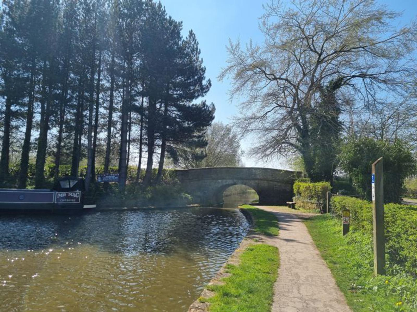 A serene canal scene shows a narrowboat moored along the left bank, with a rustic stone bridge arching over the canal. Tall pine trees line the left side, while a lush green path on the right winds toward the bridge under a clear blue sky.