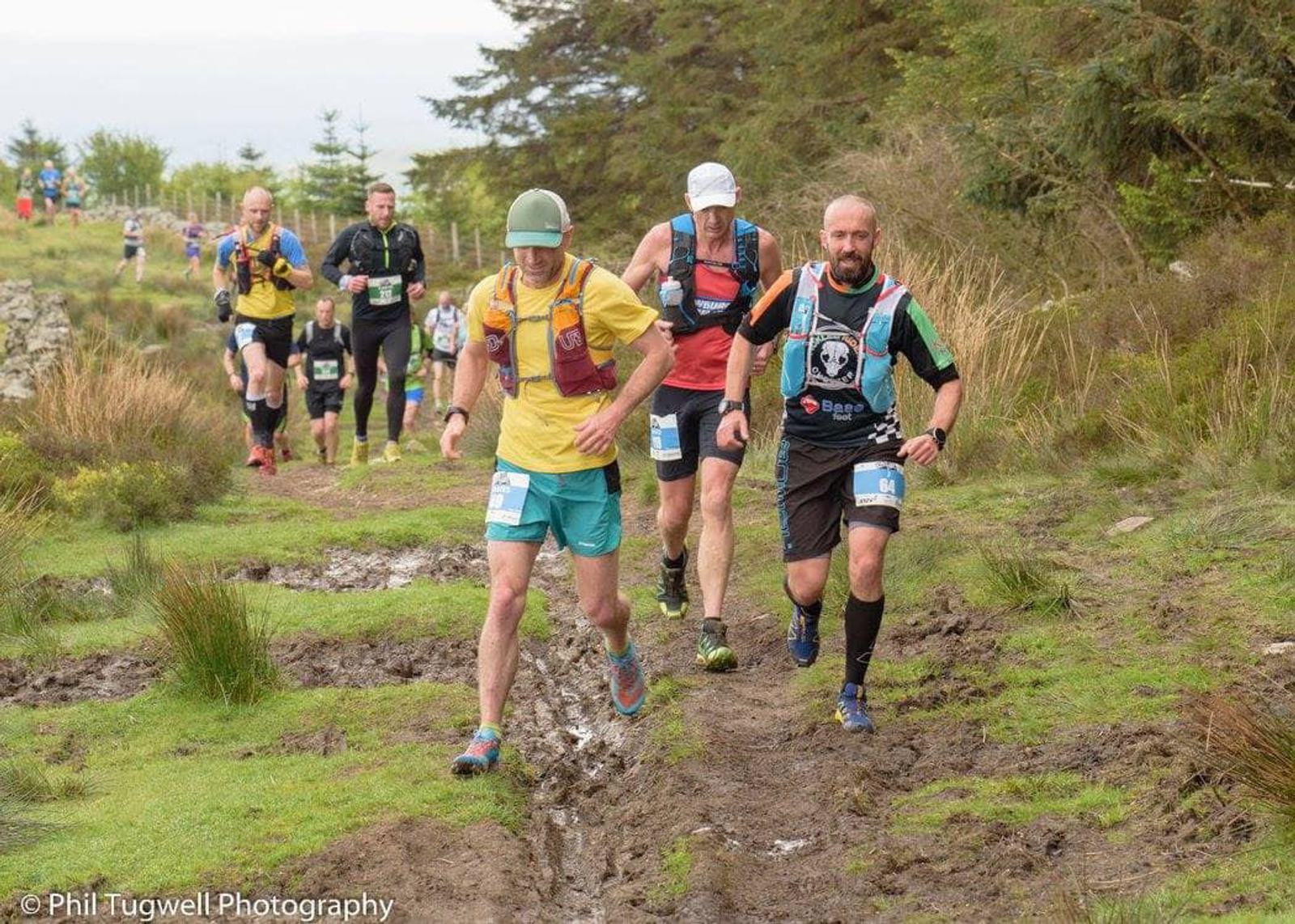 A group of runners in athletic gear navigate a muddy trail surrounded by trees and grass. They wear numbered bibs and carry hydration packs, participating in an outdoor race. The image captures the energetic atmosphere of the event.
