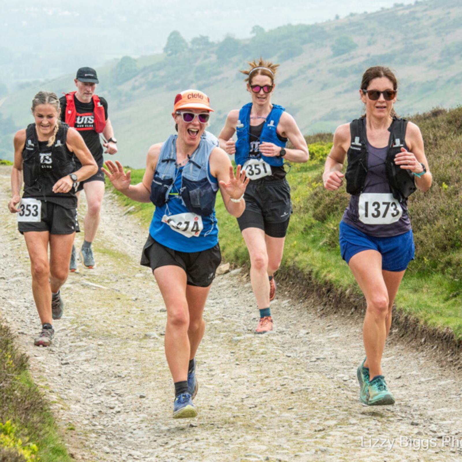 Five runners are on a trail path during a run. They're wearing outdoor athletic gear and numbered bibs. The terrain is hilly and the weather appears overcast. The mood seems energetic and focused.