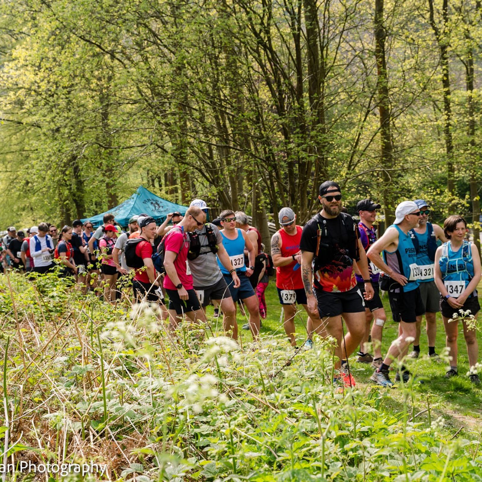 A large group of runners in colorful athletic gear stand in line on a grassy trail surrounded by trees. It's a bright day with a blue canopy visible in the background. Some runners wear race bibs with numbers.