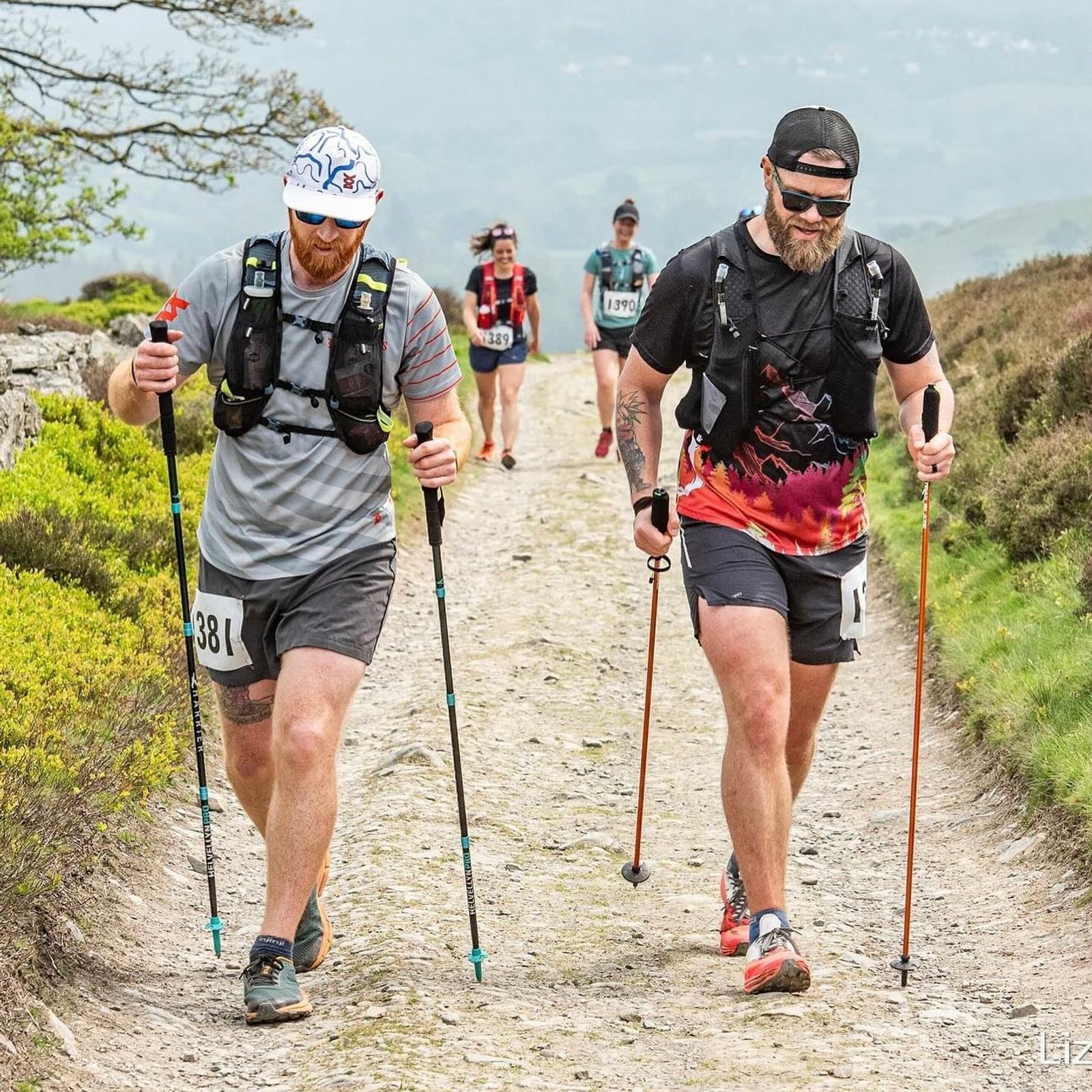 Two men are hiking on a rocky trail using trekking poles. They're wearing numbered race bibs and are accompanied by other hikers in the background. The scenery includes greenery and hills under a cloudy sky.