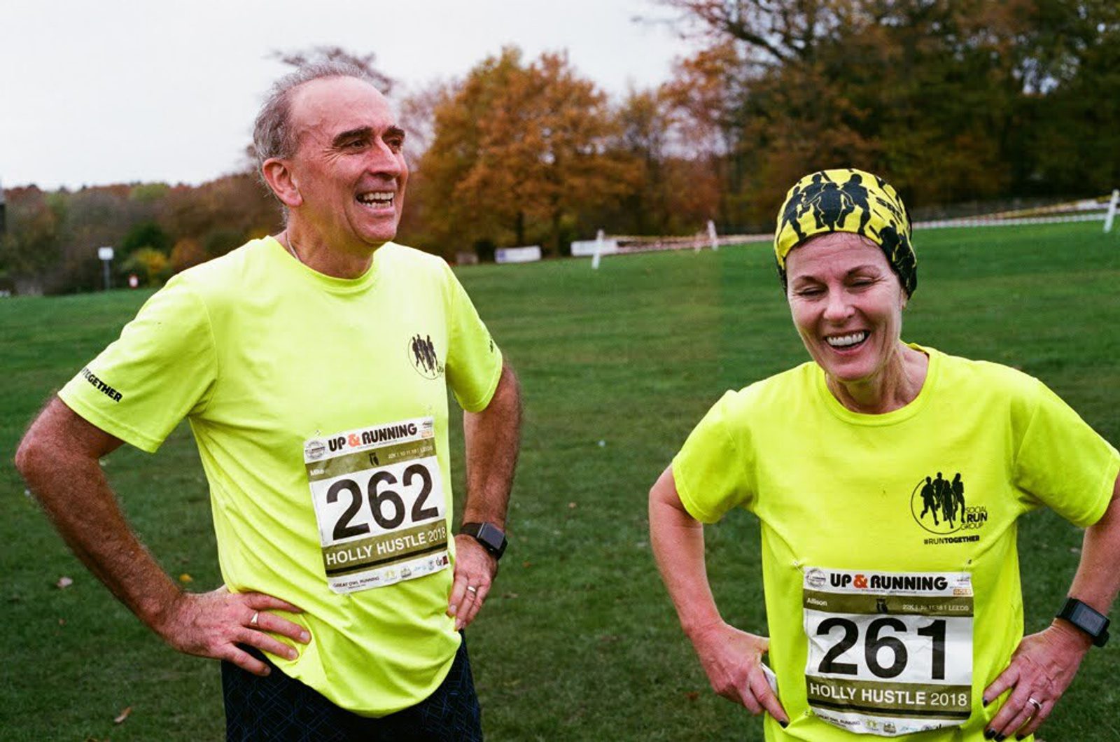Two smiling runners wearing bright yellow shirts and race bibs, numbered 262 and 261, stand on a grassy field. Trees with autumn foliage are in the background.