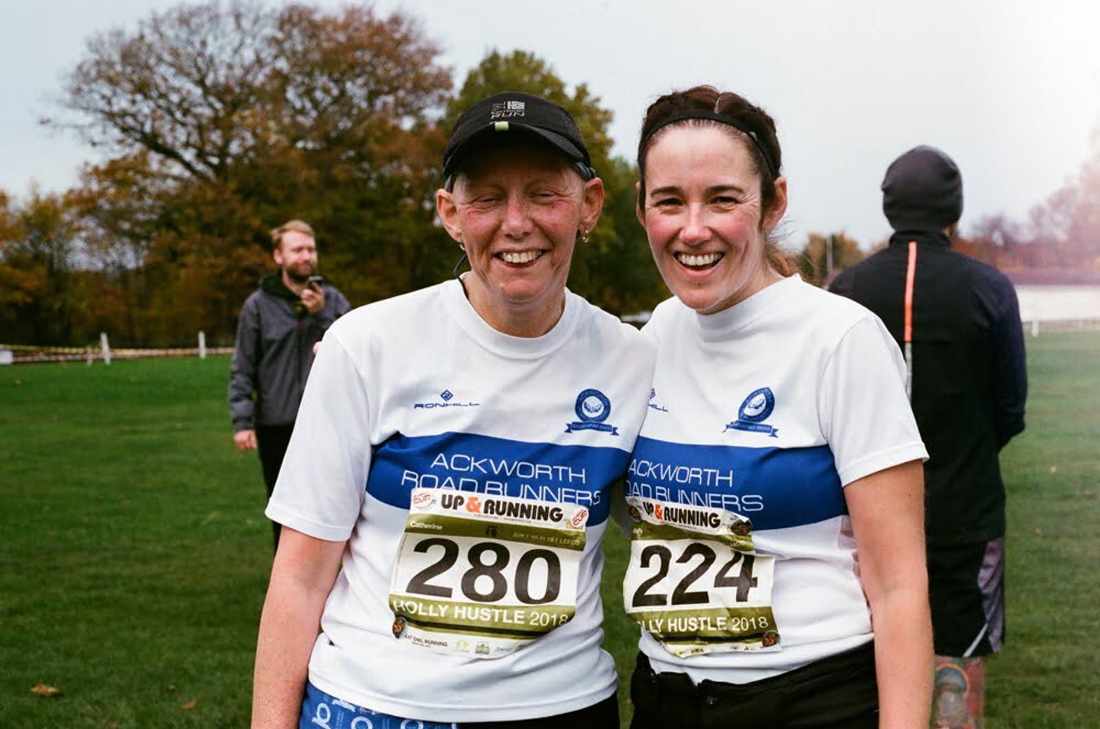 Two smiling women in matching running shirts pose together with race numbers 280 and 224. They stand on a grassy field with other participants and trees in the background.