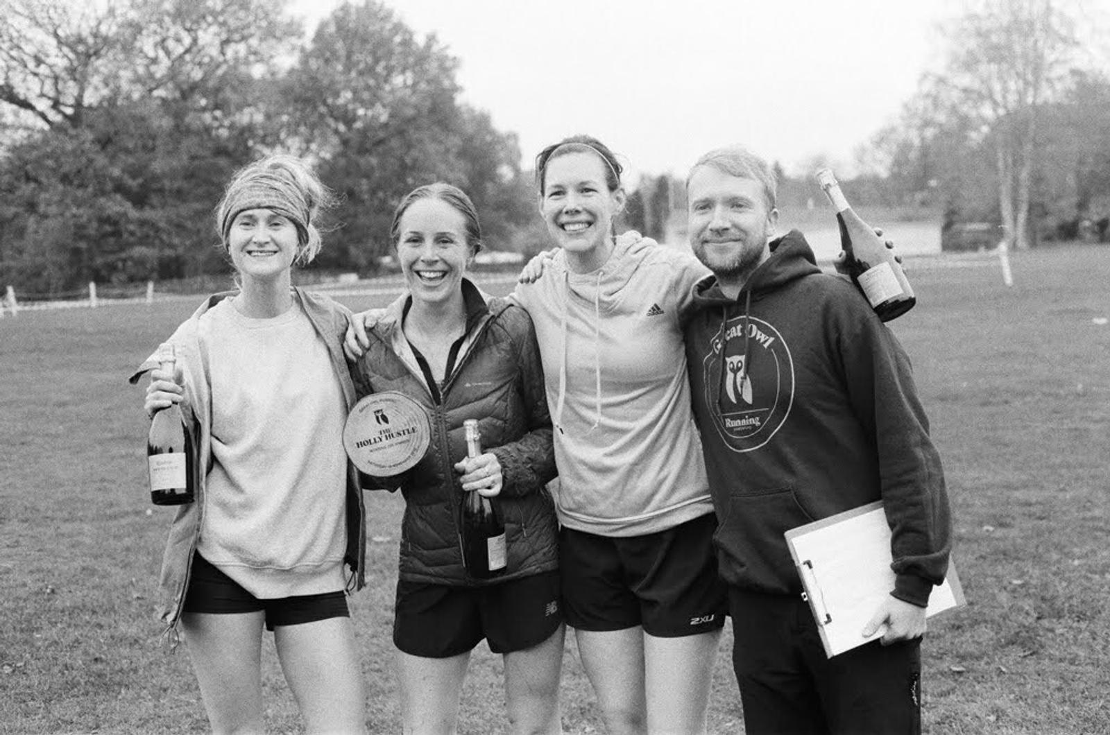 Four smiling people stand outdoors on grass, celebrating. Three hold bottles, while one person holds a plaque. They wear casual athletic clothing and pose closely with arms around each other. Trees and a fence are visible in the background.