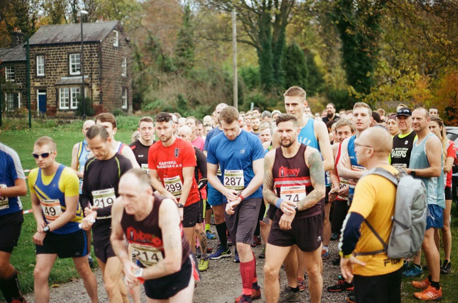 A group of runners at the starting line of a race, wearing various athletic gear and numbered bibs. Trees and two stone houses are visible in the background on a cloudy day.
