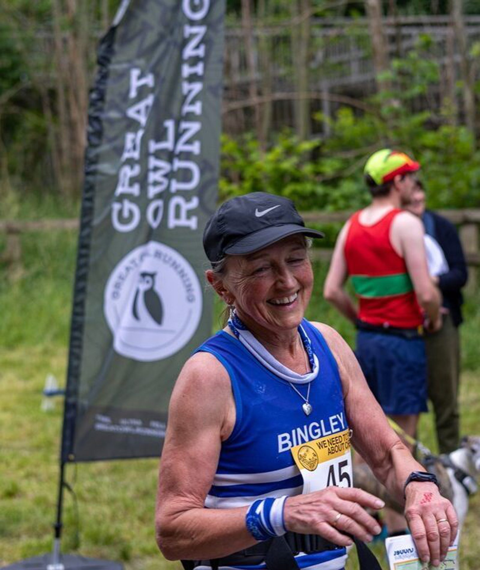 A smiling runner wearing a blue and white "Bingley" jersey approaches the finish line, with other participants in the background. A banner reading "Great Owl Running" stands nearby. A circular sign on the left reads "We Need to Talk About Chevin.