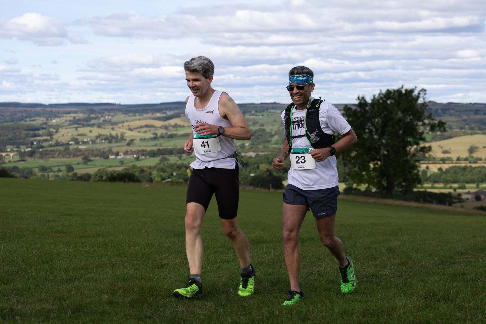 Two runners on a grassy hill wearing race bibs numbered 41 and 23. They are in athletic gear, smiling, with a scenic countryside landscape in the background under a partly cloudy sky.