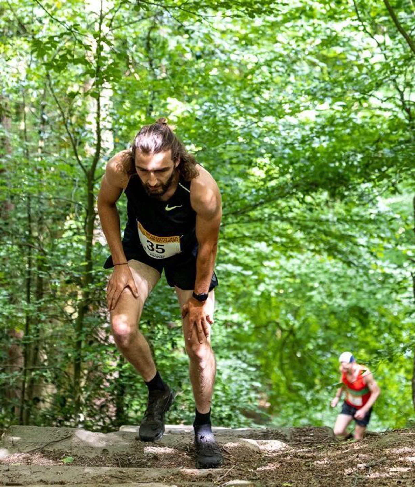A runner in a black tank top and shorts leans forward while ascending a steep forest trail. Another runner in a red shirt is visible in the background. A circular sign reads "We Need to Talk About Chevin." Sunlight filters through the trees.