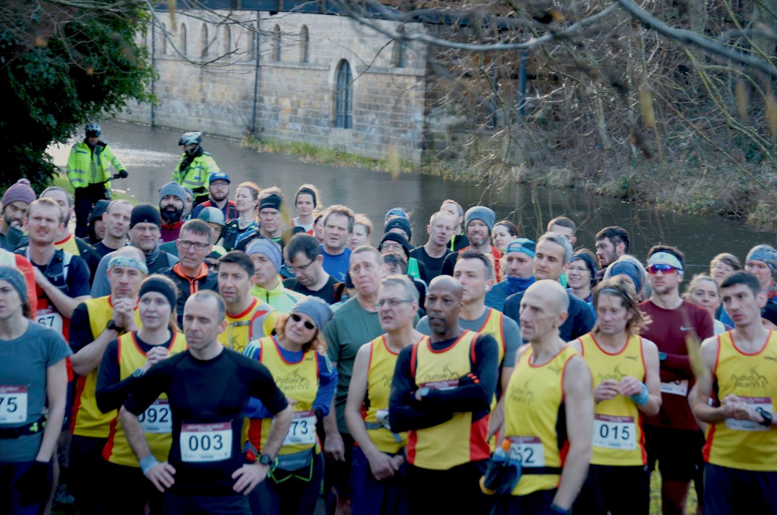 A large group of people wearing running gear gather at the start of a race near a river. They're dressed in various colors with race numbers. Two police officers on bikes are visible in the background near a stone bridge. It's an overcast day.