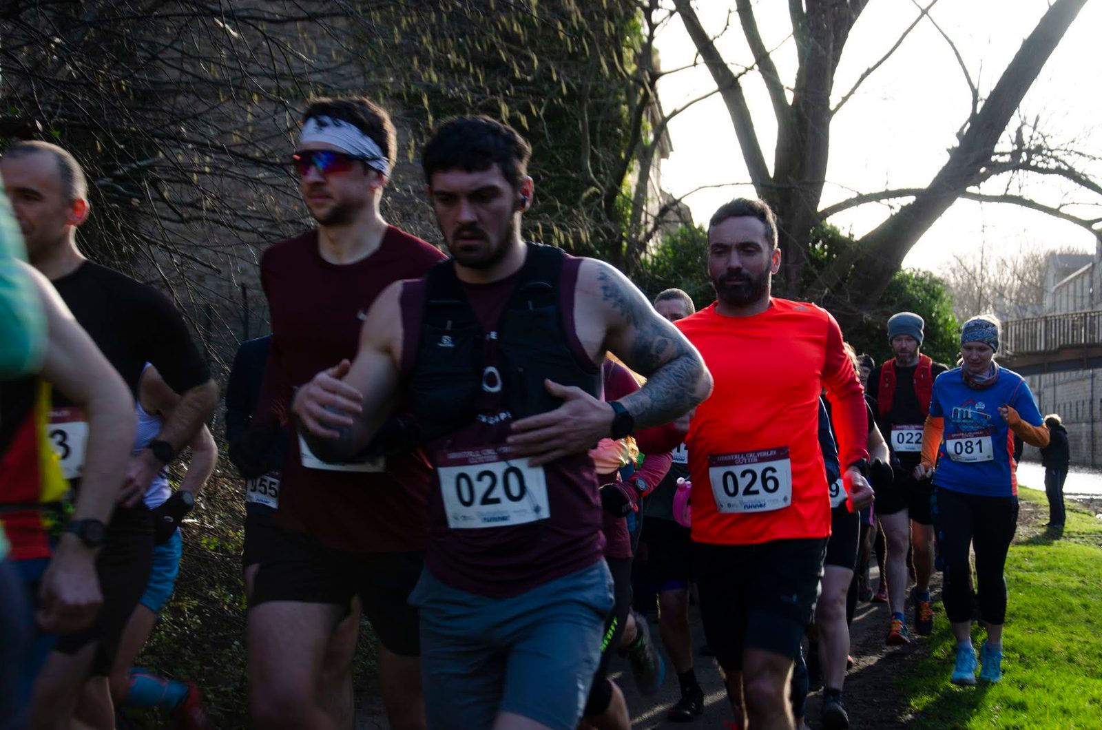 A group of runners participating in an outdoor race, wearing numbered bibs. The runners are dressed in athletic gear, including tank tops and long sleeves. Trees and natural surroundings are visible in the background.