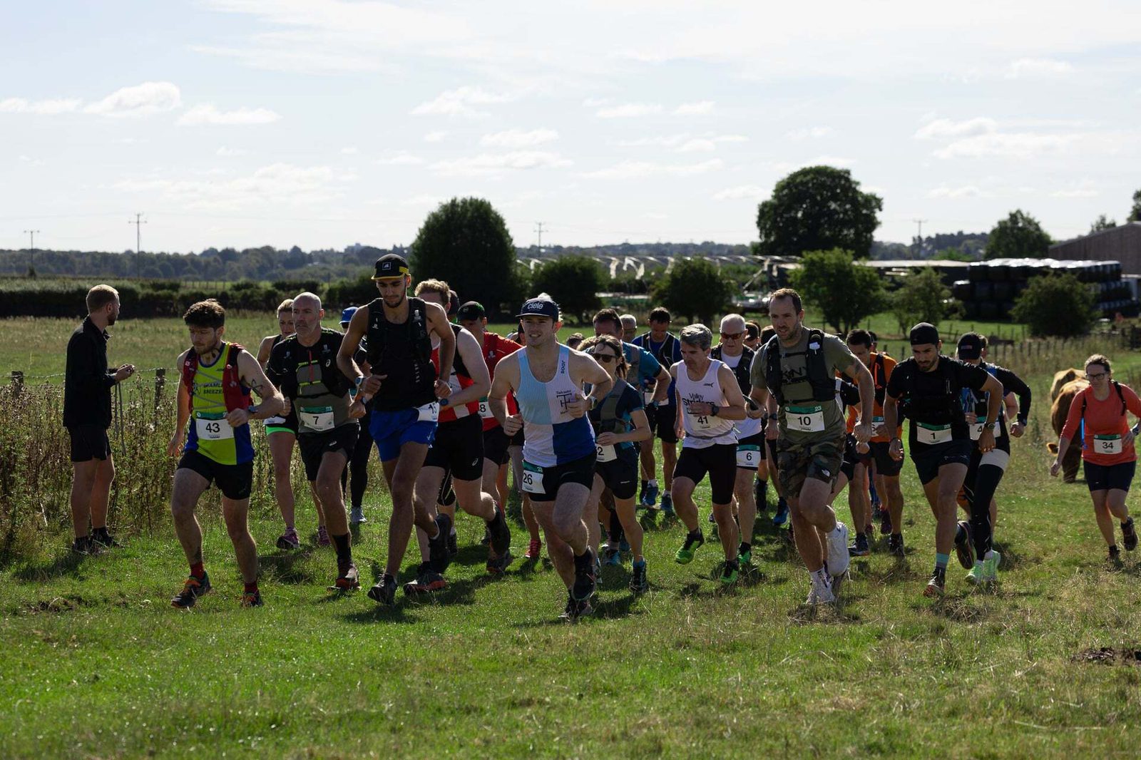 A group of runners begins a race on a grassy field under a sunny sky. Participants wear bib numbers and athletic gear, with some trees and a scenic backdrop visible in the distance.
