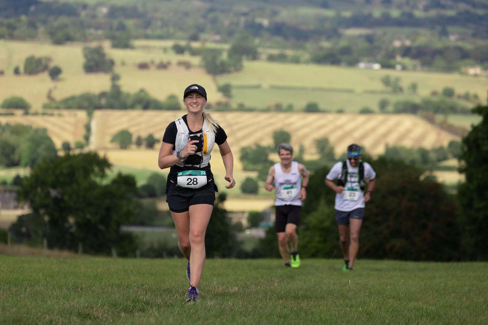 A woman in athletic gear and a cap, wearing bib number 28, runs through a scenic countryside. She's smiling as two other runners follow behind. The landscape features rolling hills and fields under a cloudy sky.