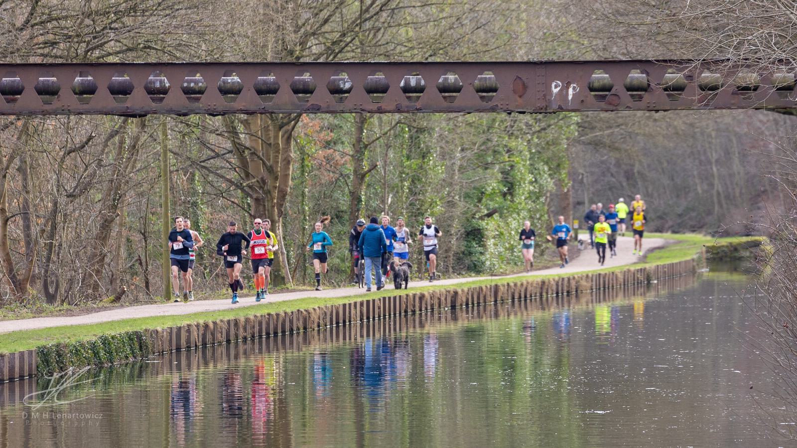 Runners and walkers on a path beside a canal, with a rusty metal bridge overhead. The scene is surrounded by bare winter trees, and reflections are visible in the water.