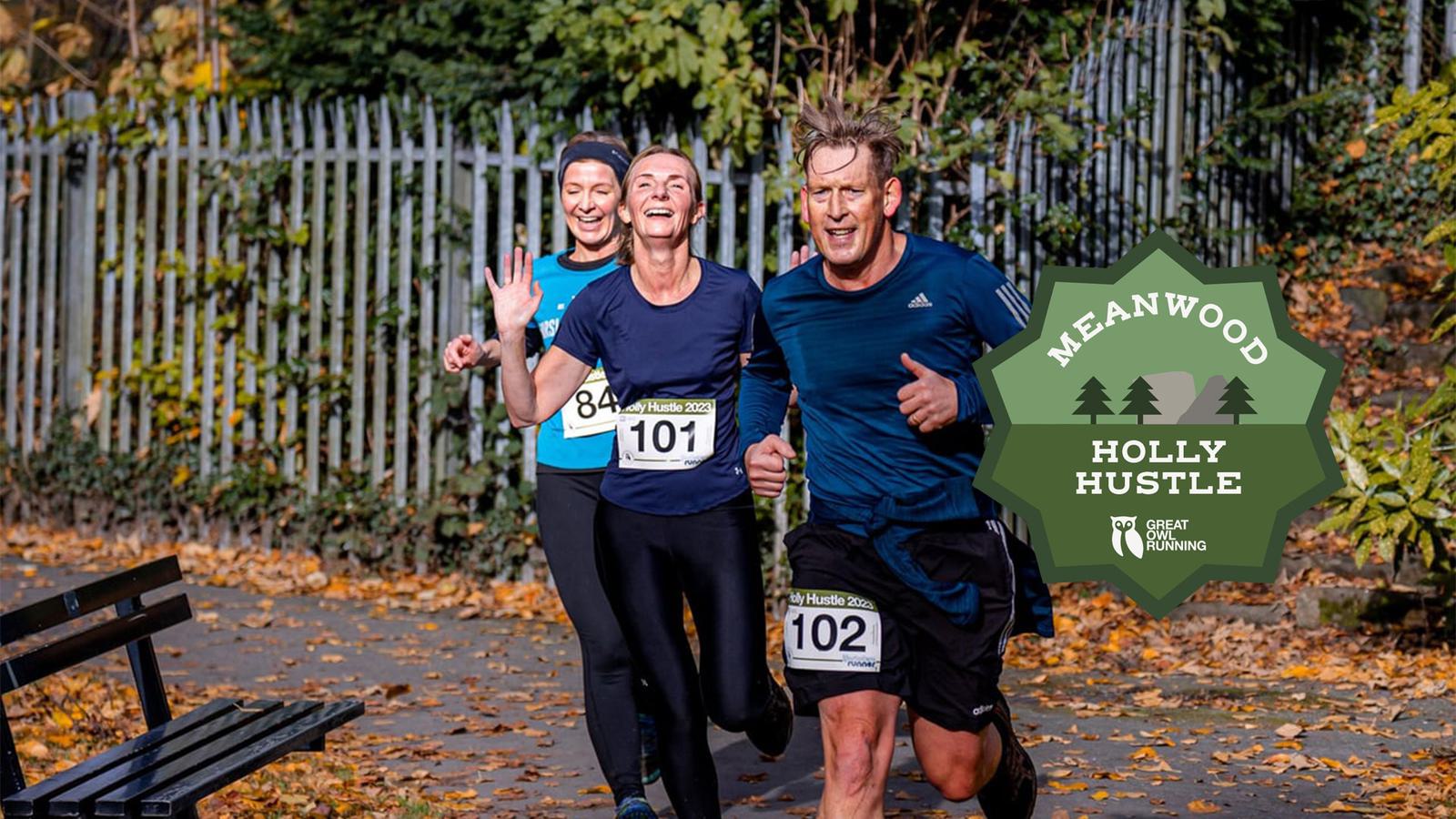 Three runners with race numbers 84, 101, and 102 are smiling and jogging in a park during the Meanwood Holly Hustle event. They are surrounded by autumn leaves, and a green event logo is visible.