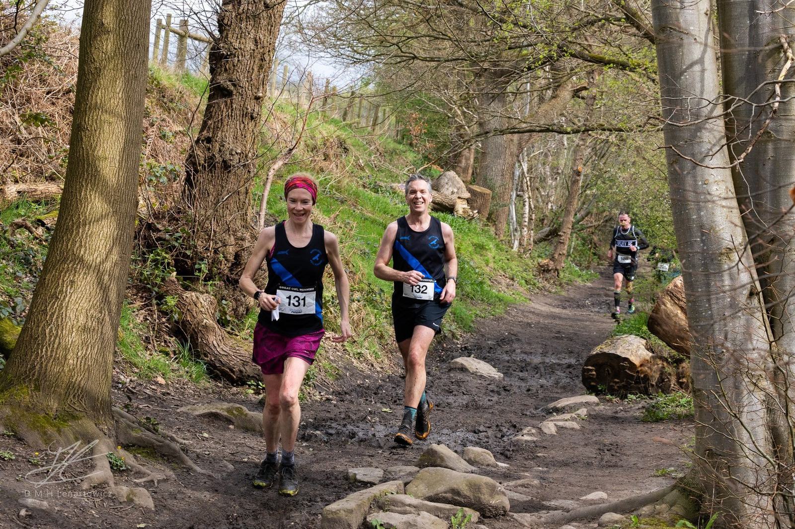 Three runners on a forest trail race, wearing numbered bibs. The path is rocky and surrounded by trees. It's a sunny day, and they appear focused and determined.