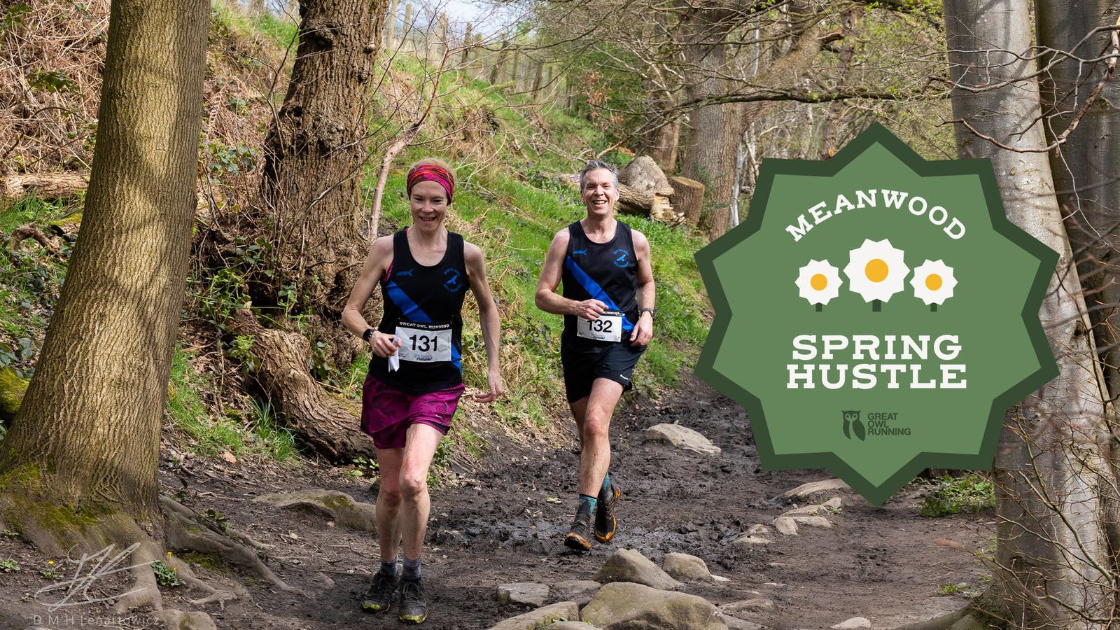 Two runners in a forest trail race, wearing numbered jerseys 131 and 132. They are smiling and running on a rocky path surrounded by trees. A sign reads "Meanwood Spring Hustle" with white flowers.