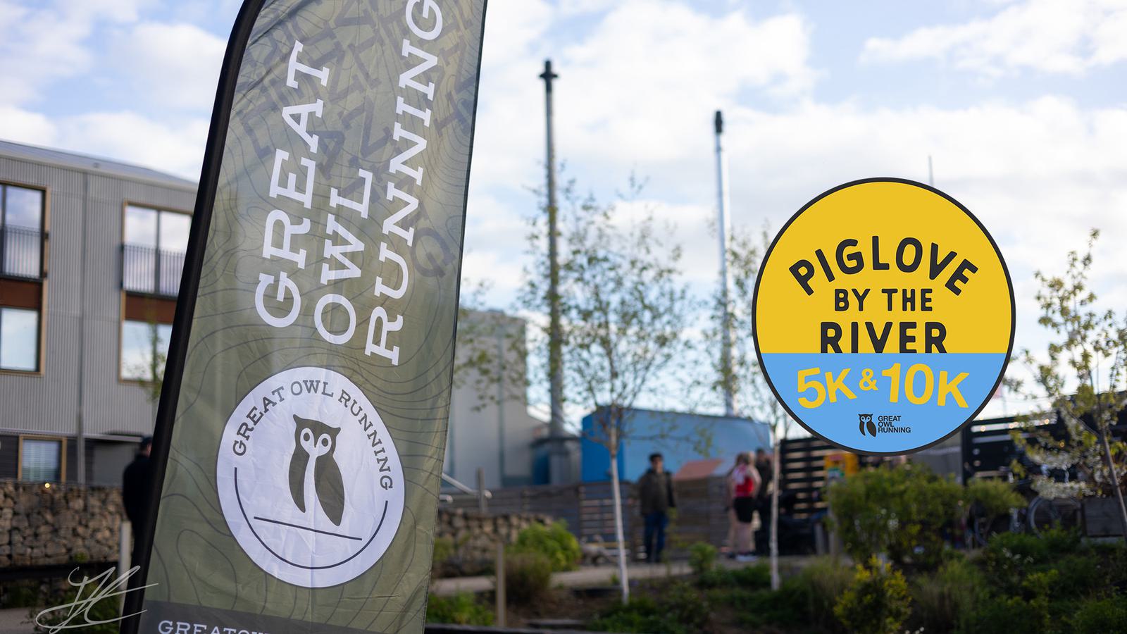 A banner with "Great Owl Running" logo next to a circular sign reading "Piglove by the River 5K & 10K." Blurry background showing trees and a building.