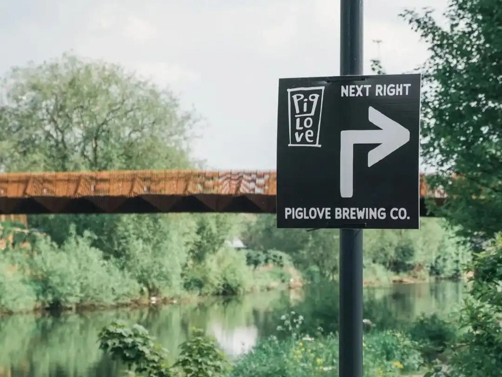 A sign indicating "Piglove Brewing Co. Next Right" with a right-pointing arrow. In the background, there is a lush green landscape and a brown bridge over a river.