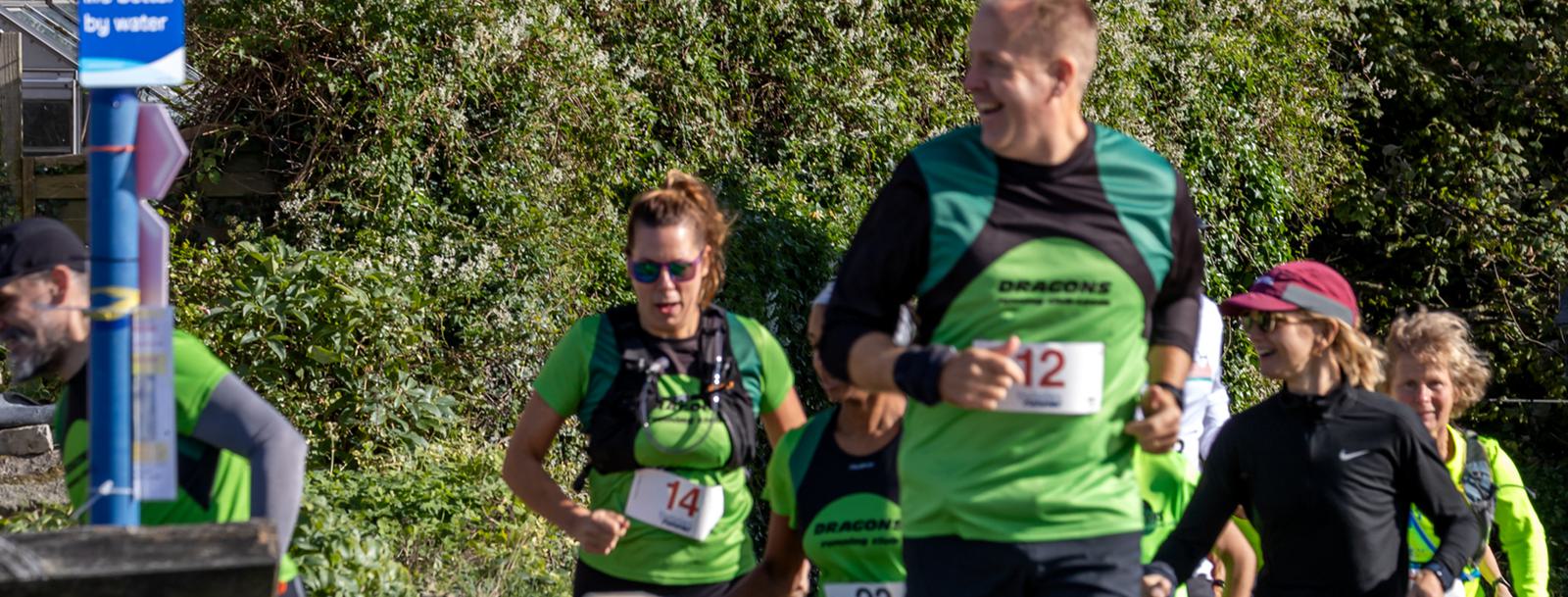 A group of runners in green shirts with race numbers, participating in an outdoor event on a sunny day. They're running along a path surrounded by greenery. Some runners wear sunglasses and caps. The atmosphere appears lively and energetic.