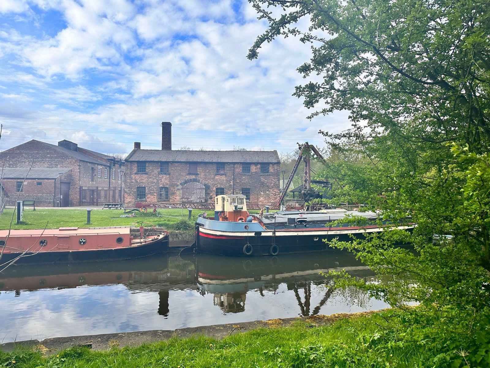 A tranquil scene of a historic brick building by a canal, featuring two docked boats and lush greenery. The sky is partly cloudy, adding to the serene atmosphere.