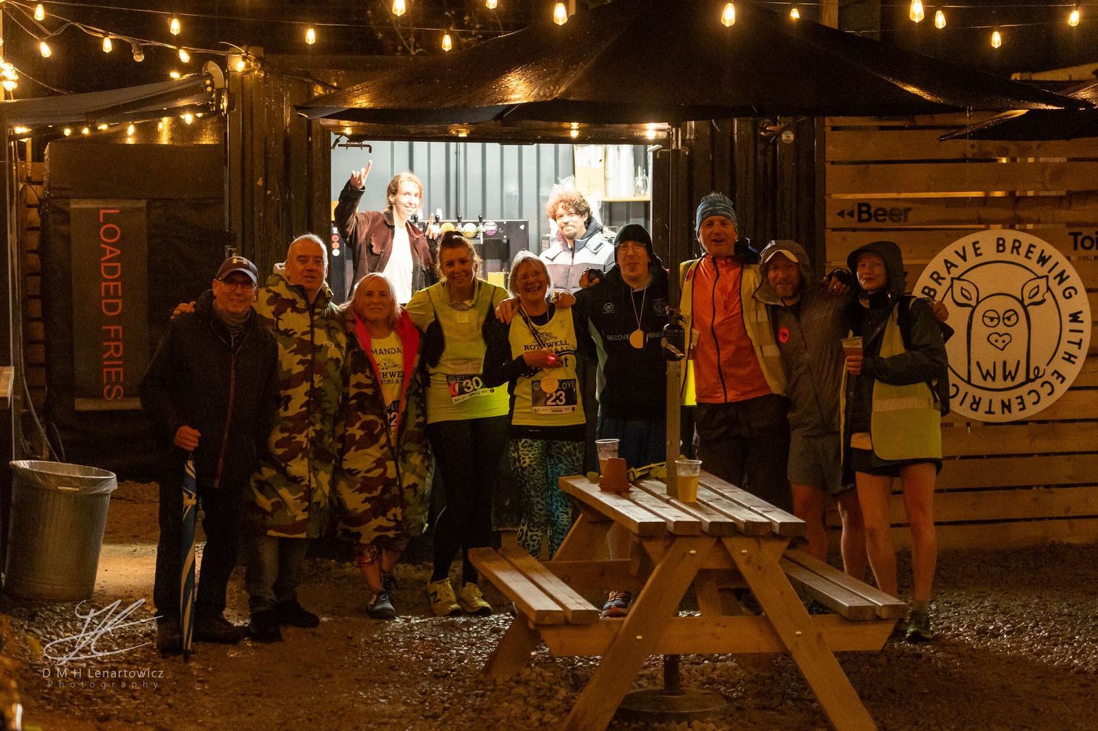 A group of people posing happily under string lights at night near a wooden bench. Some wear running gear and high-visibility vests. A woman in the back is pouring drinks inside a container bar that has a sign reading "Brave Brewing.