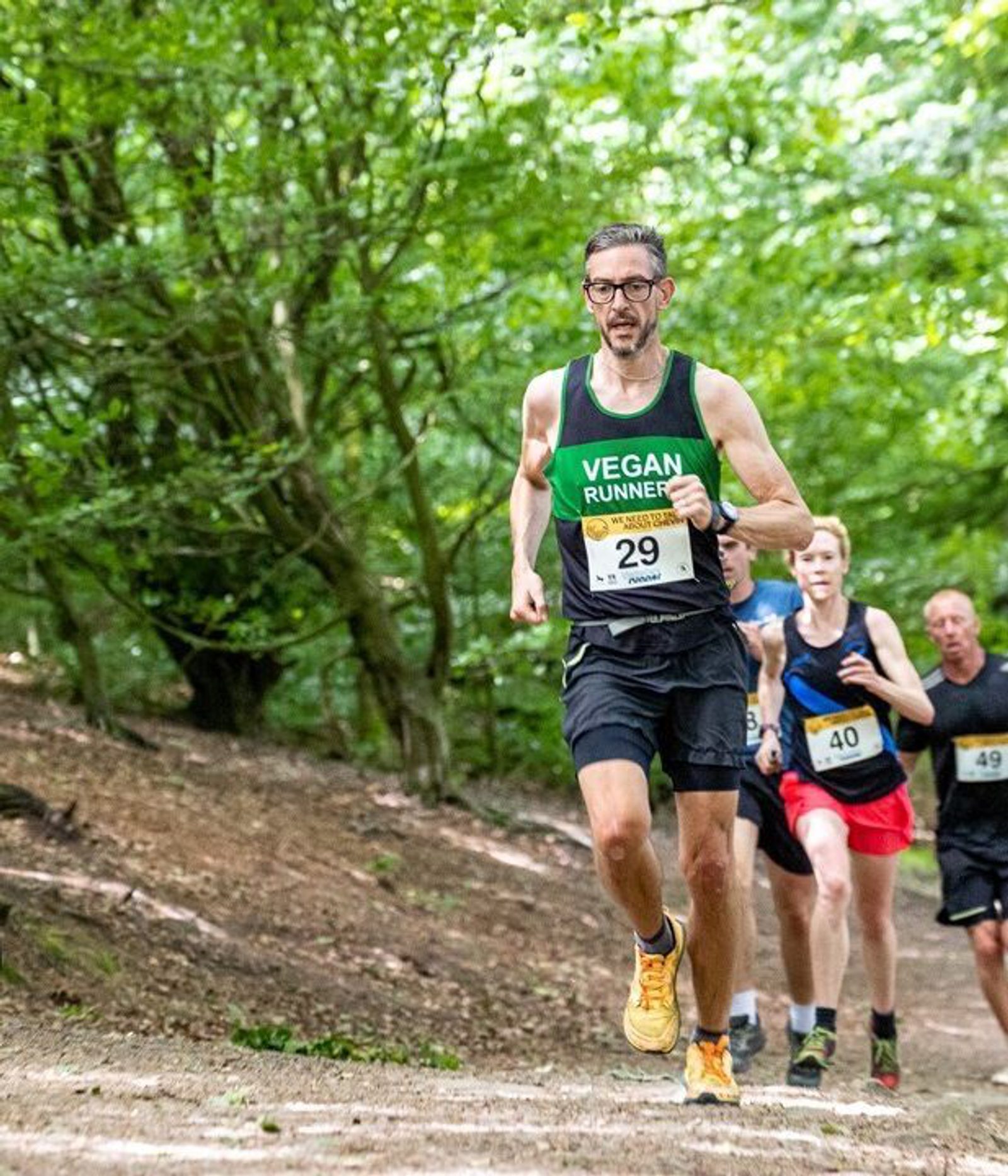 A runner in a green "Vegan Runner" tank top runs through a wooded trail. Two other runners follow closely behind. The trail is surrounded by lush greenery. A round sign says, "We need to talk about Chevin.