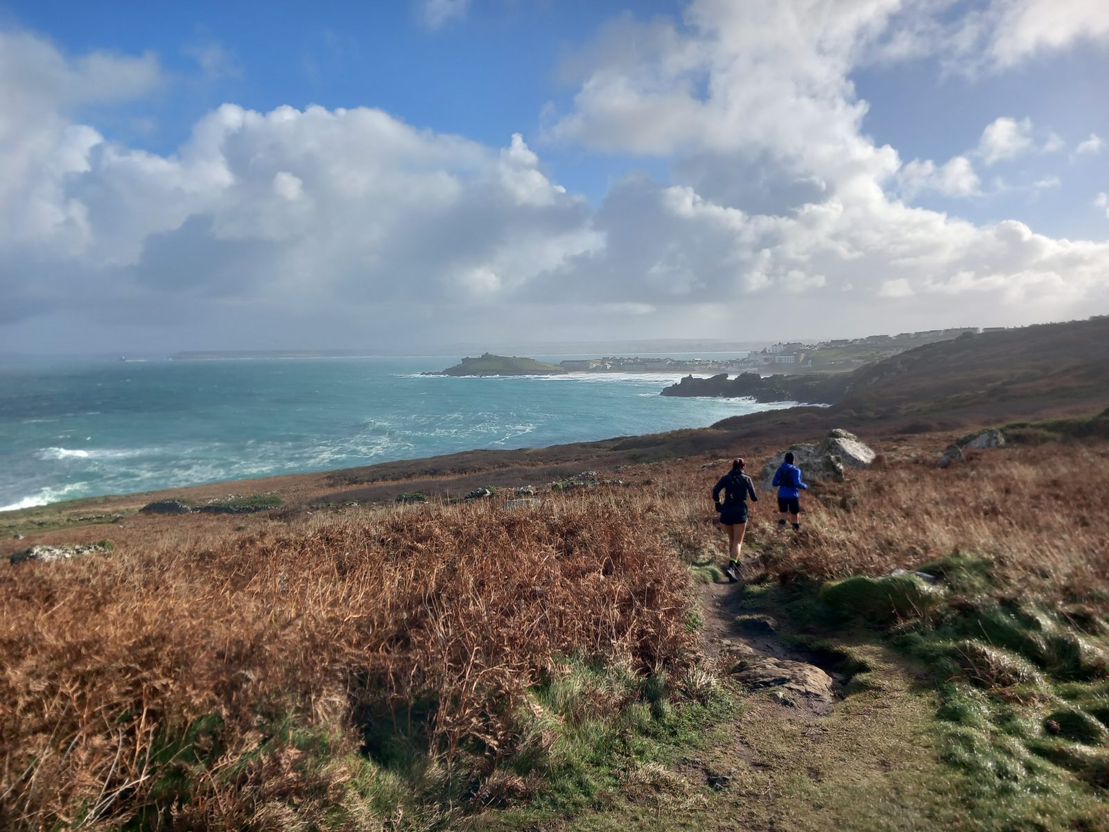 Two people are jogging along a coastal path. The scene shows a large expanse of rugged terrain with dry grass and a narrow trail. To the left, the ocean meets the coast, with waves crashing against the shore. The sky is cloudy but bright, indicating a breezy day.